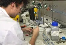 Scientist in a lab coat is pictured injecting something into a small glass vial on a lab bench